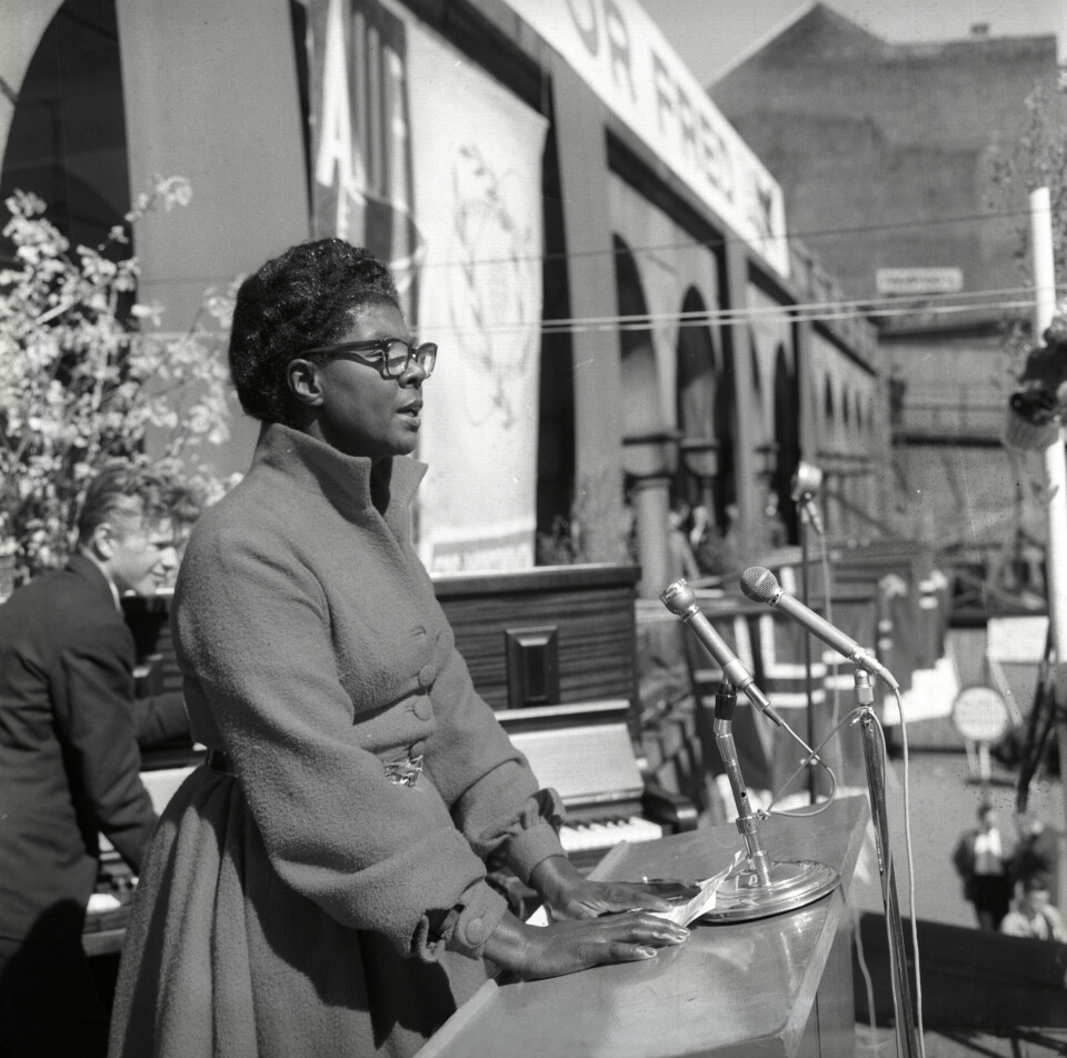 Et sort-hvitt bilde som viser en kvinne som ser ut som hun holder en tale, Hun har flere mikrofoner foran seg.