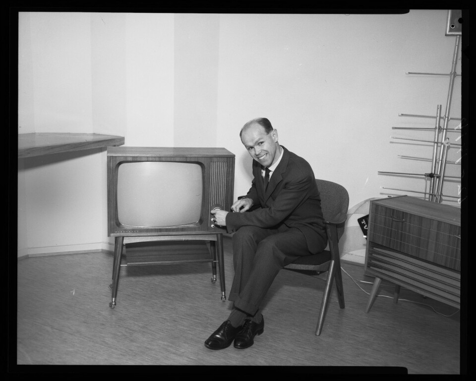 Foto til tv-reklame Nordmende 1961. Sort-hvitt-bildet viser en smilende mann i dress som sitter i en stol ved sien av en tv på bein