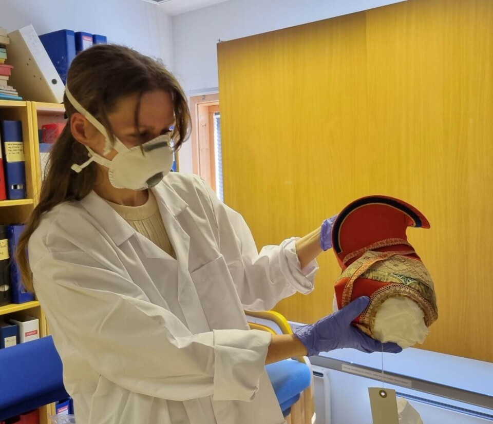 Ingvild Marie Bjørnå Pettersen, tidligere leder av Várjjat Sámi Musea / Varanger Samiske Museum, pakker opp Bååstede gjenstandene. Bildet viser en kvinne med langt hår. Hun har på seg en hvit frakk, munnbind og blå hansker og holder en gjenstand i glade farger.