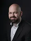 Portrett av Kjetil Landrog i mørk jakke og hvit skjorte mot svart bakgrunn.