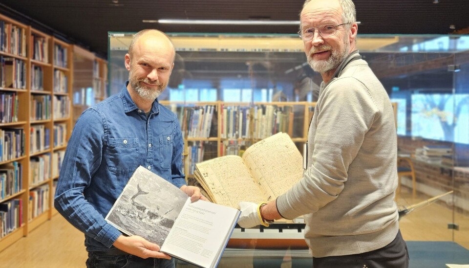 Seksjonsleder ved Norsk folkemuseum Ole Myhre Hansen (t.h) og Yngve Schulstad Kristensen (t.v) arkivar ved KUBEN fotografert på Norsk Maritimt Museum under overrekkelsen kaptein Crawfurds dagbok i oktober 2024. To menn holder hver sin protokoll og ser mot kameraet. De står i et rom med hyller med med bøker langs veggene
