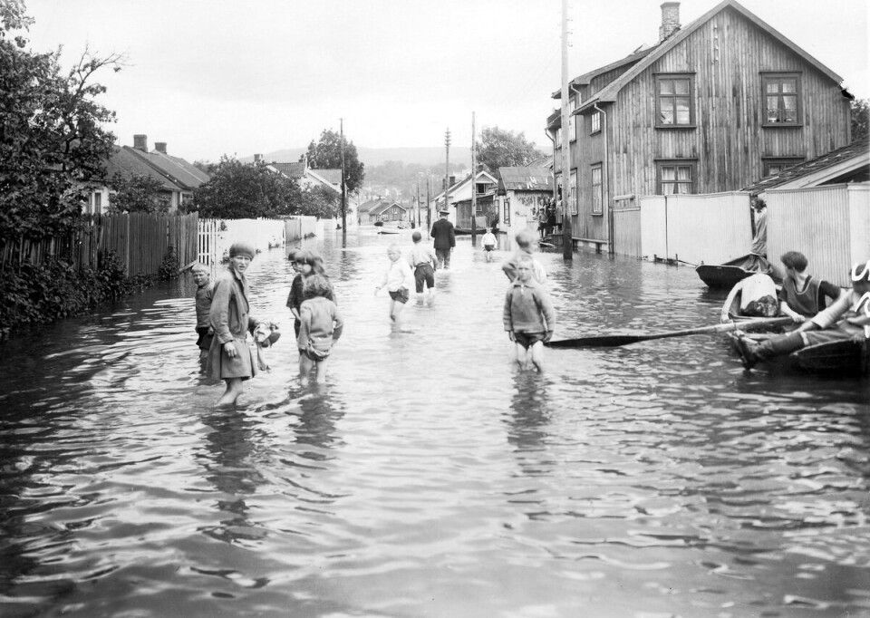 SKIEN 1927: Bildet er tatt under flommen i Skien i 1927. Sort-hvit-bilde som viser en gate fylt med vann. Det er trehus og gjerder på begge sider, og folk både vasser og bruker robåt.