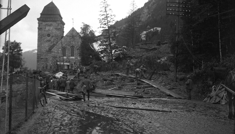 RJUKAN 1927: Oppryddingsarbeid etter ras ved Rjukan kirke i 1927. Mennesker lager gangveier i rasområdet med klopper. Hus er rast sammen og ligger inntil kirkens nordvegg. Sort-hvit-bildet viser en kirke i bakgrunnen og en rasert og våt eller gjørmete vei foran.