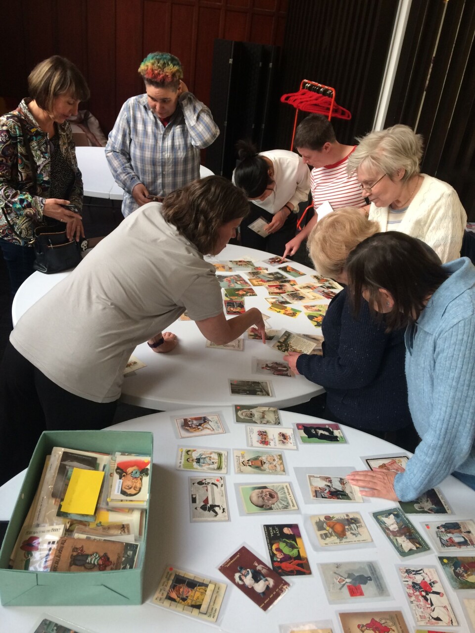 ÅPENT ARKIV: Glasgow women´s library har en stor samling propagandapostkort fra begge sider i kampen om kvinners stemmerett på tidlig 1900-tall. Mange er både sjokkerende og groteske og gir innblikk i datidens holdninger. Her fra et arrangement i 2023. Bildet viser en gruppe med sju kvinner som står lent over et bord fylt av postkort.