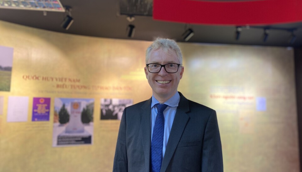 Arkivverkets Ole Gausdal (Arkivverket) er programansvarlig for Nordiske arkivdager 2025. Bildet viser en blond mann med briller, dress og slips. Han står foran en gylden vegg med noen bilder/plakater på.