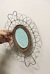 Hånd holder ovalt veggoppheng med turkis blomsterrelieff foran lys vegg