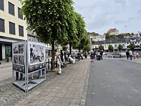 Utendørsutstilling med fotoplakater langs en gate i Arendal sentrum, med folk som går forbi.