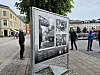 Stort utendørs skilt med historiske svart-hvite bilder på et torg med noen personer i nærheten.