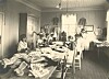 Sort-hvitt-bilde. Flere kvinner sitter rundt et langt bord med tekstiler og papir i et lyst arbeidsrom.