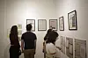 Tre personer ser på innrammede kunstverk og informasjonstavler på en vegg i et galleri.