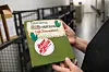 Hender som holder en grønn bok med dekorert omslag og håndskrevet tekst.