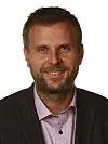 Smilende skjeggete mann i mørk jakke og rosa skjorte foran hvit bakgrunn.