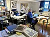 Person ved et skrivebord i et lyst arbeidsrom med bøker, papirer og kunst på veggene.