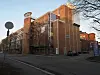 Stor rødbrun mursteinsbygning ved en gate, med trær, biler og blå himmel.